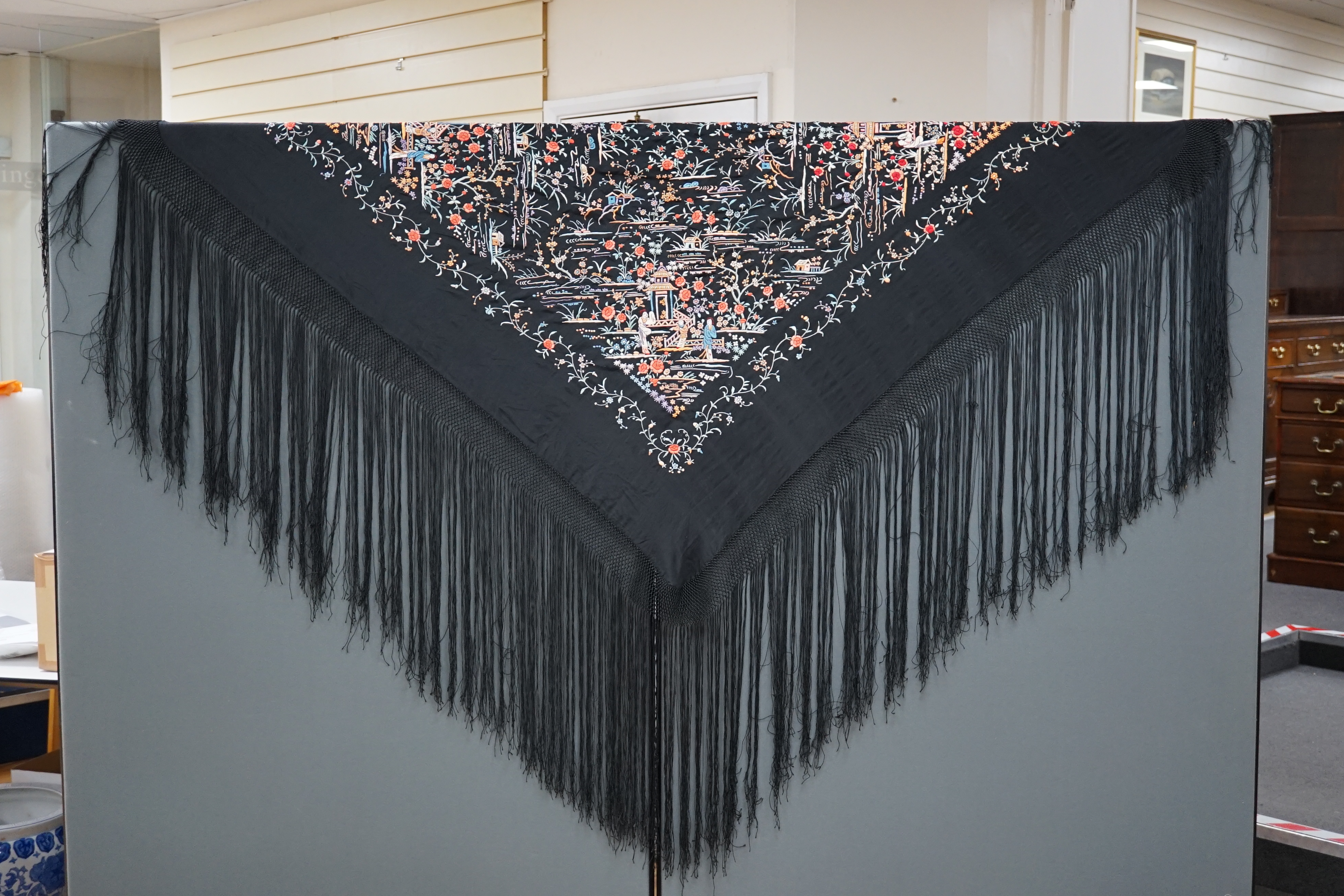 An early 20th century Chinese black silk shawl, with all over coloured floral embroidery, embroidered with multi-coloured silks and a long silk fringe, unusually the four corners are embroidered with a figurative pagoda